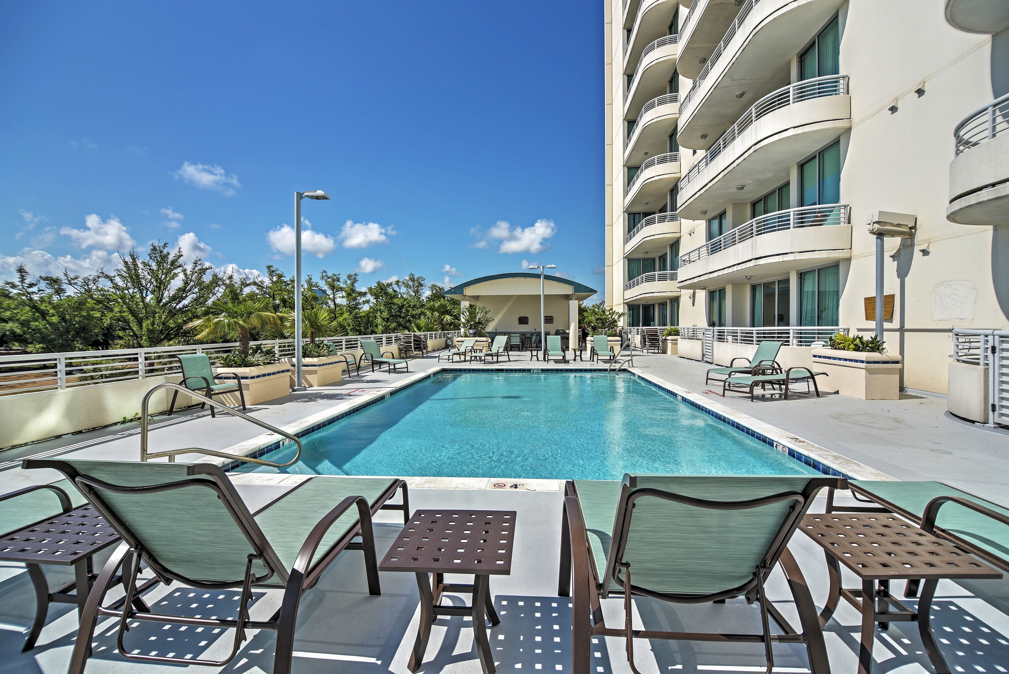 A swimming pool with lounge chairs and tables, a building with balconies, and a clear blue sky.