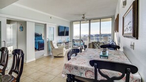 A spacious living area with a dining table, chairs, and a view of the city through large windows. - Beachside Biloxi Club Condo: Balcony w/ Ocean View (Biloxi)