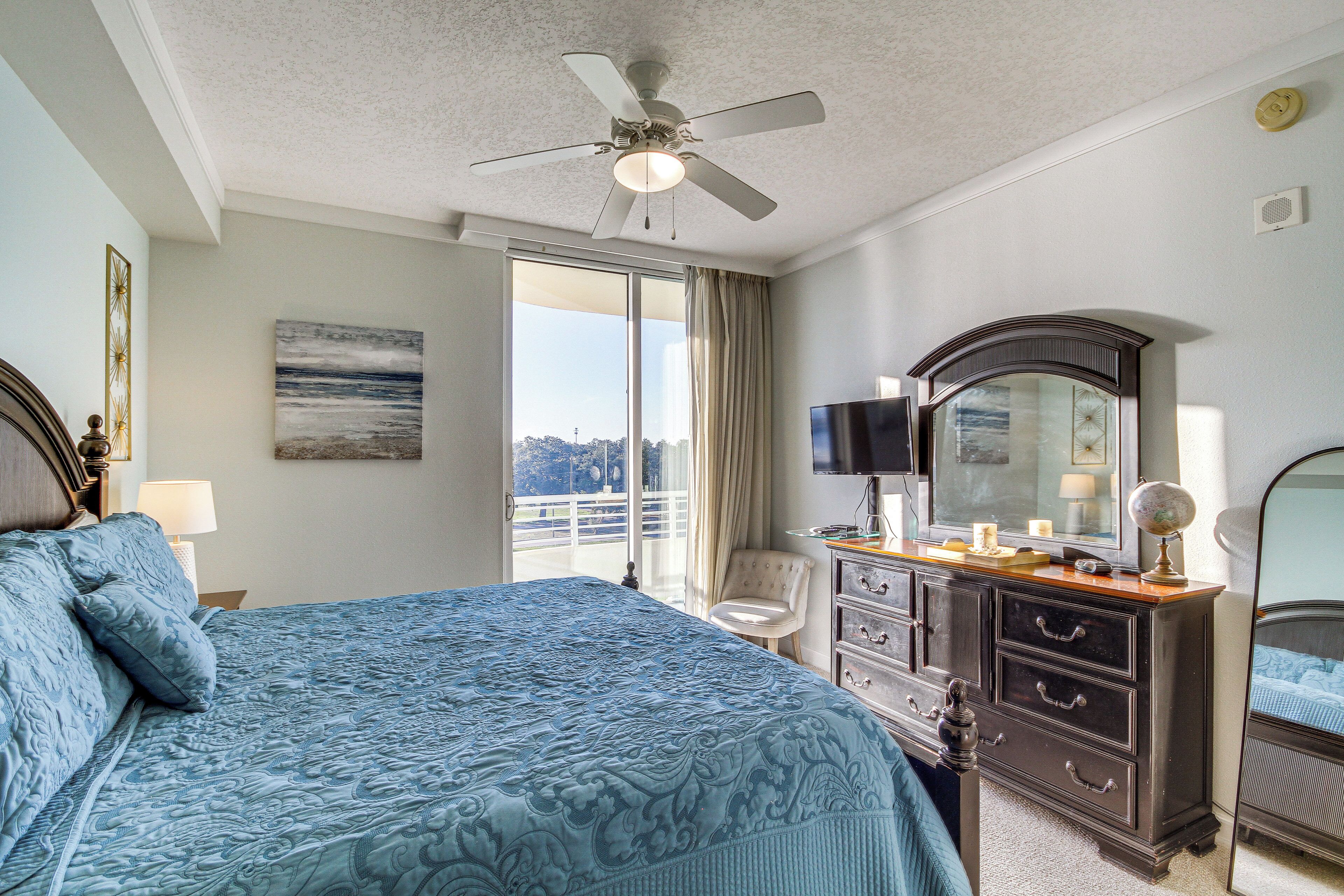 A bedroom with a large bed, a ceiling fan, a dresser with drawers, a television, and a large window with curtains.