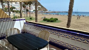 Une terrasse extérieure avec une table et des chaises, donnant sur une plage où l’on peut voir des gens et des bateaux.