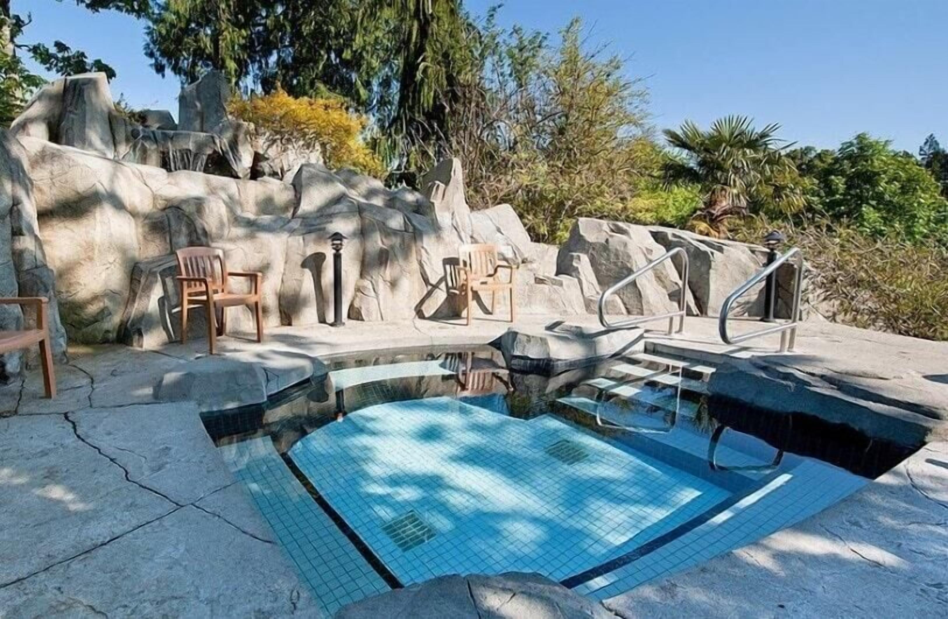 A swimming pool with a rock wall backdrop, surrounded by chairs and a sunshade.