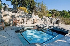 Piscina com parede de rocha ao fundo, rodeada por cadeiras e um toldo.