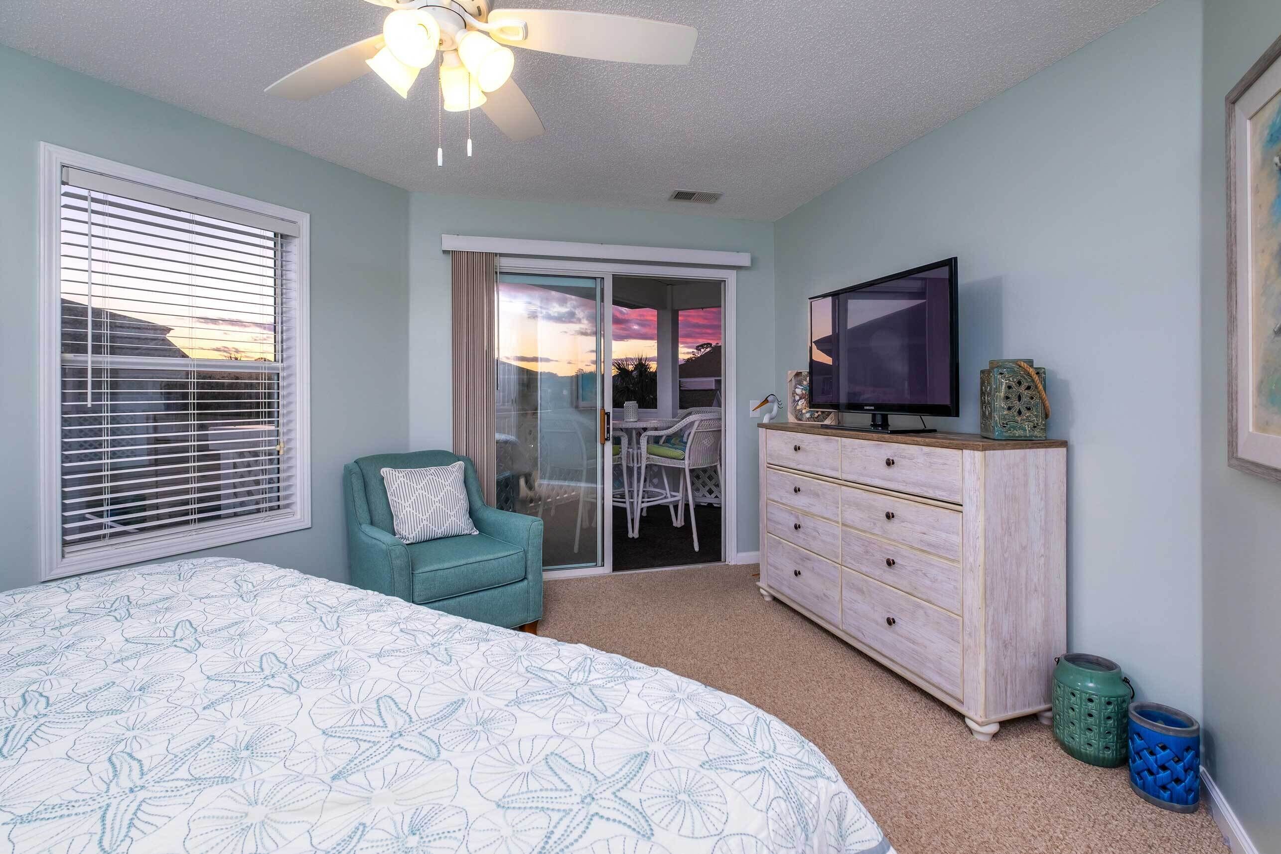 A bedroom with a bed, a TV, a dresser, a chair, and a sliding glass door leading to a balcony.