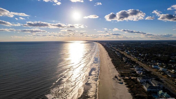 Vue aérienne d’une plage avec une longue étendue de sable et une rangée de maisons le long de la côte.