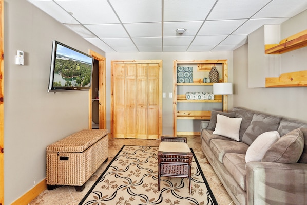 A living room with a sofa, a television mounted on the wall, a wooden wardrobe, a wicker bench, and a patterned rug.