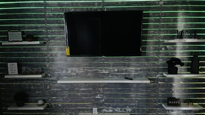 A modern interior with a flat-screen TV mounted on a wooden wall with green LED lighting. There are shelves with various items and a small sign that says "Welcome to our home." - Hidden Gem Downtown Florence - Near UNA (Florence)