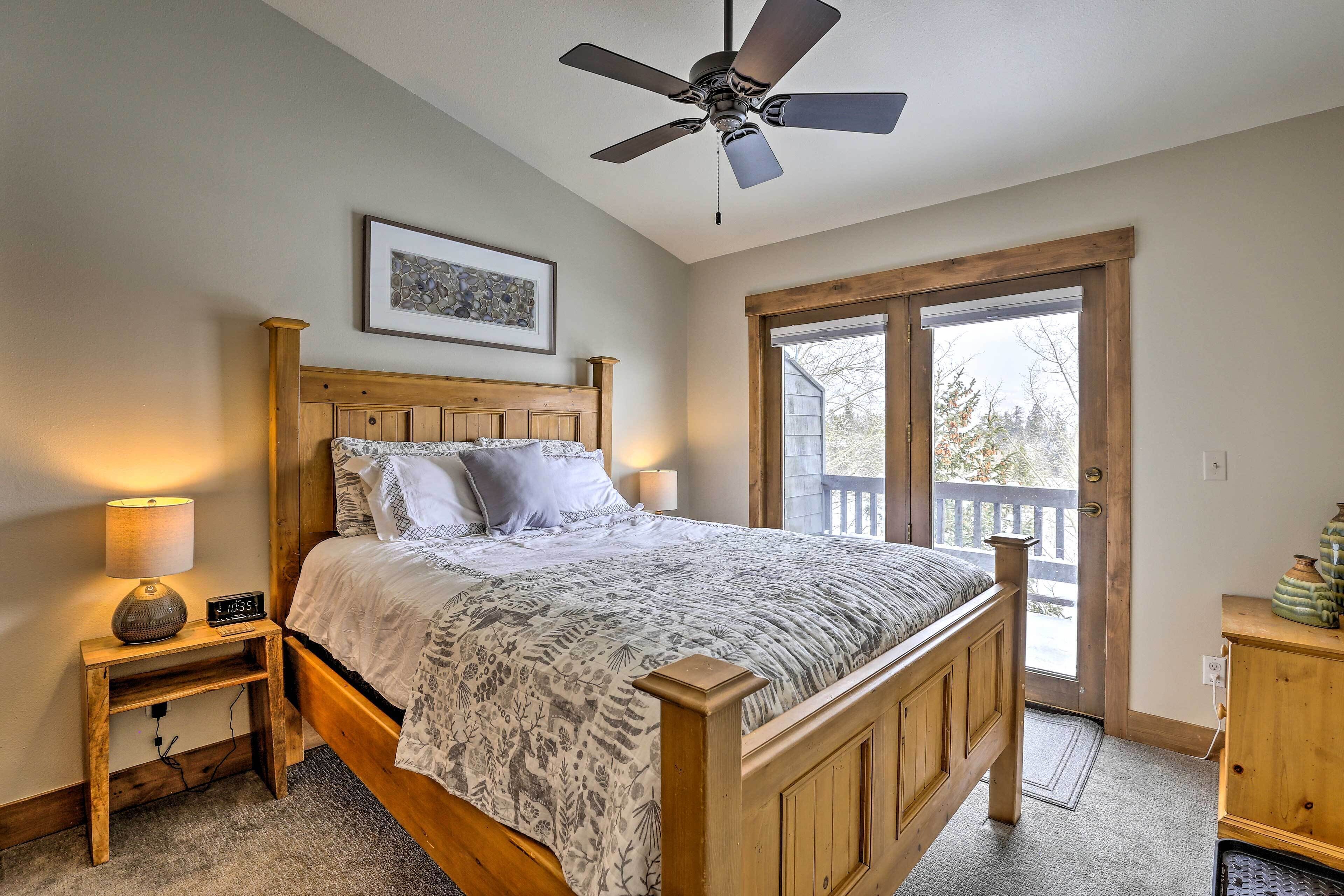 A bedroom with a wooden bed, a ceiling fan, a nightstand with a lamp, a framed picture on the wall, and a view through a door.