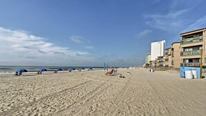 Une plage avec des parasols bleus, une rangée de chaises longues et un bâtiment doté de balcons.