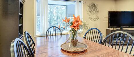A dining area with a wooden table, chairs, a vase with flowers, a television, and a window with curtains.