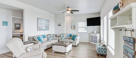 A spacious living room with a ceiling fan, a sofa set, a coffee table, and a small ottoman. There are wall-mounted shelves with decorative items and a sign on the wall.
