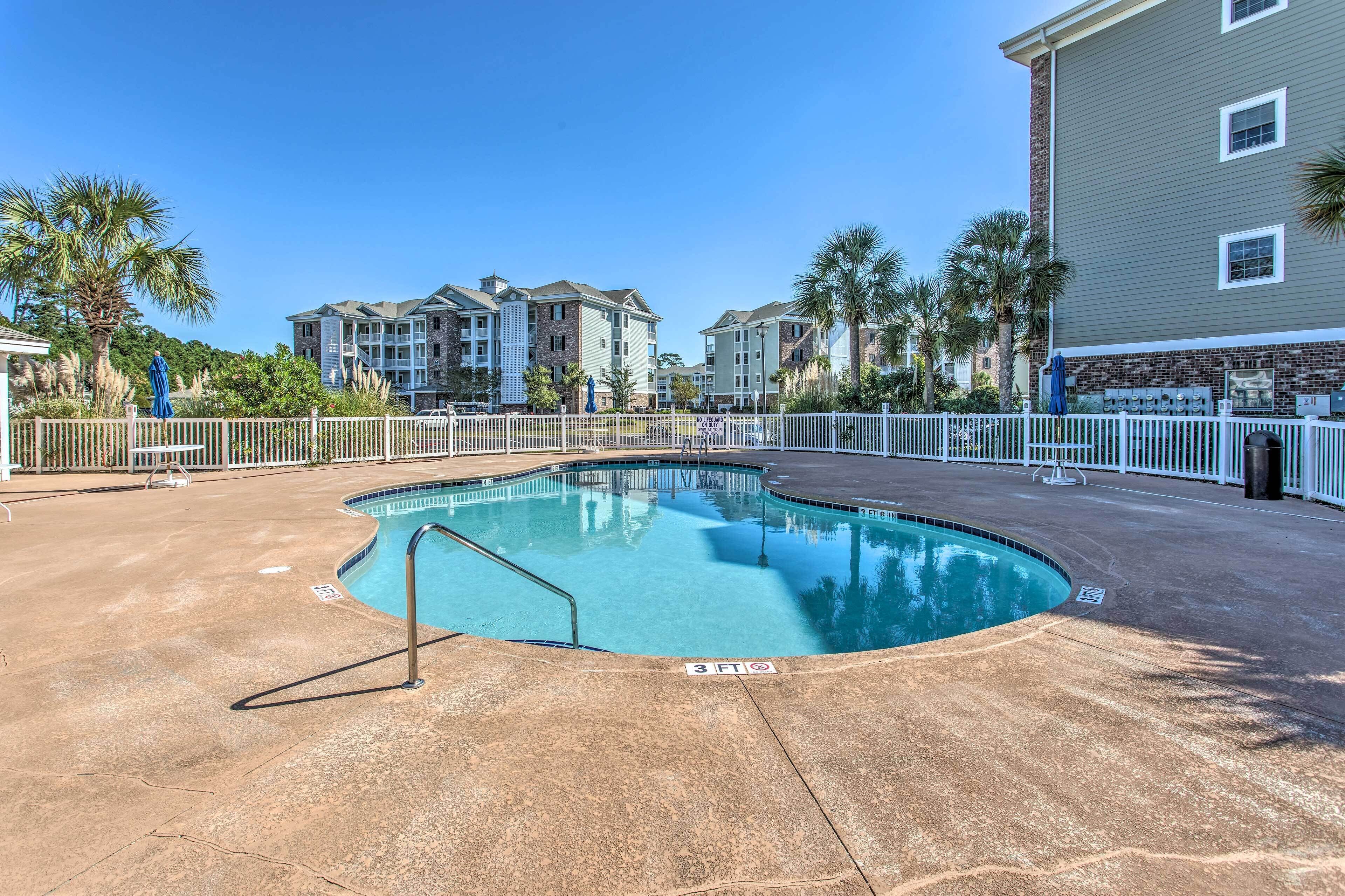 A swimming pool with a 3 ft depth marker, surrounded by a white fence, with multi-story buildings in the background.