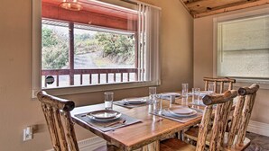 A dining area with a wooden table set for four, surrounded by rustic chairs, large windows with blinds, and a view of a road and greenery outside. - Luxe Cabin w/ Covered Pool + Smoky Mountain Vistas (Sevierville)