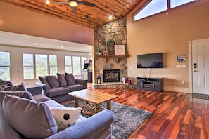 A spacious living room with a stone fireplace, a television, and a large window overlooking a scenic landscape. - Luxe Cabin w/ Covered Pool + Smoky Mountain Vistas (Sevierville)