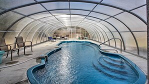 An indoor swimming pool with a curved design, surrounded by chairs and tables, under a transparent roof structure. - Luxe Cabin w/ Covered Pool + Smoky Mountain Vistas (Sevierville)