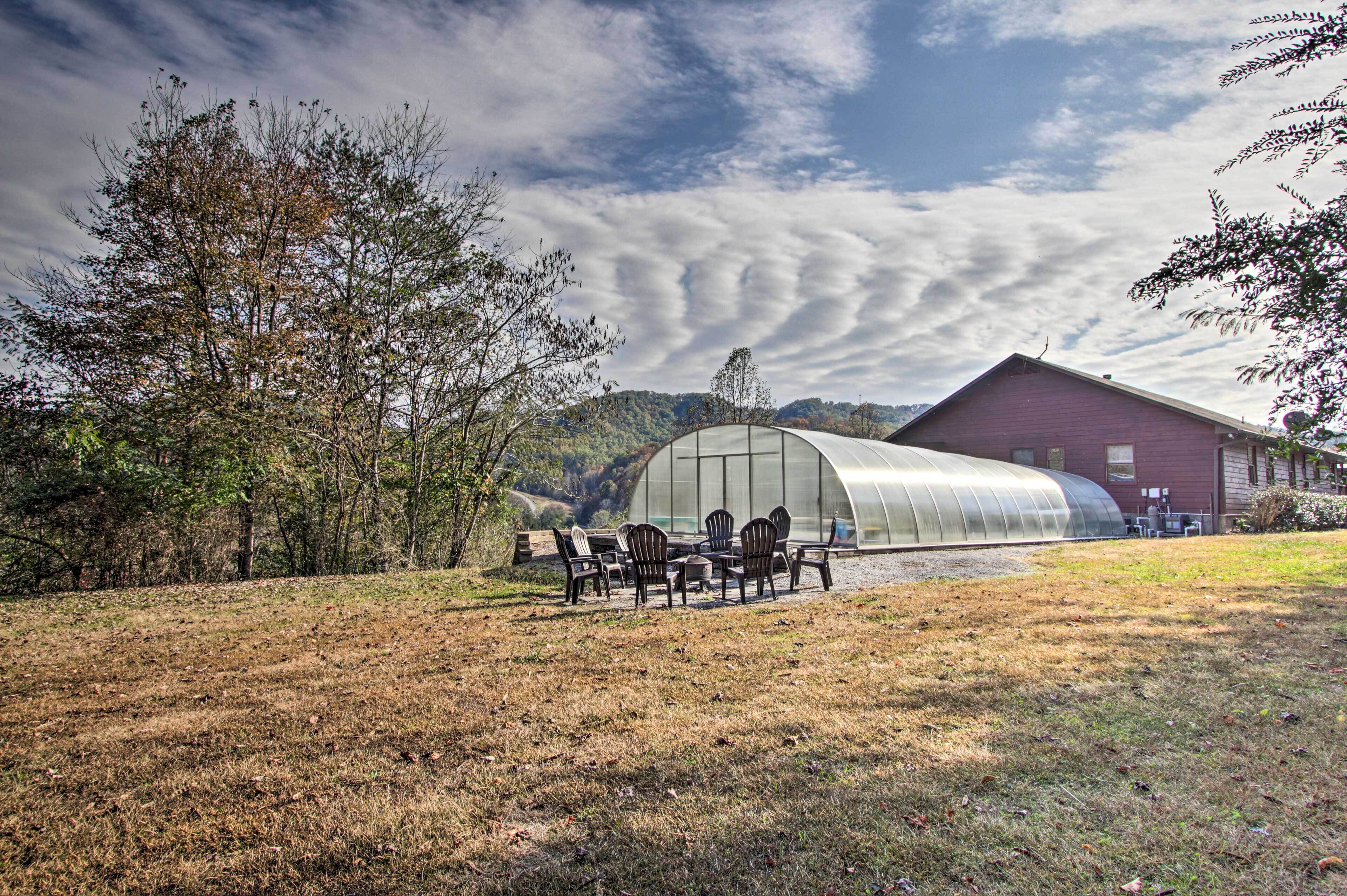 A greenhouse with a curved roof, a dining table with chairs, and a red building in the background.