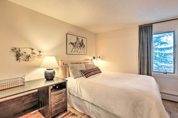 A bedroom with a bed, bedside lamps, a wall-mounted artwork, a window with curtains, and a wooden nightstand with drawers. - Breckenridge Condo w/ Gas Grill < 1 Mi to Peak 8! (Breckenridge)