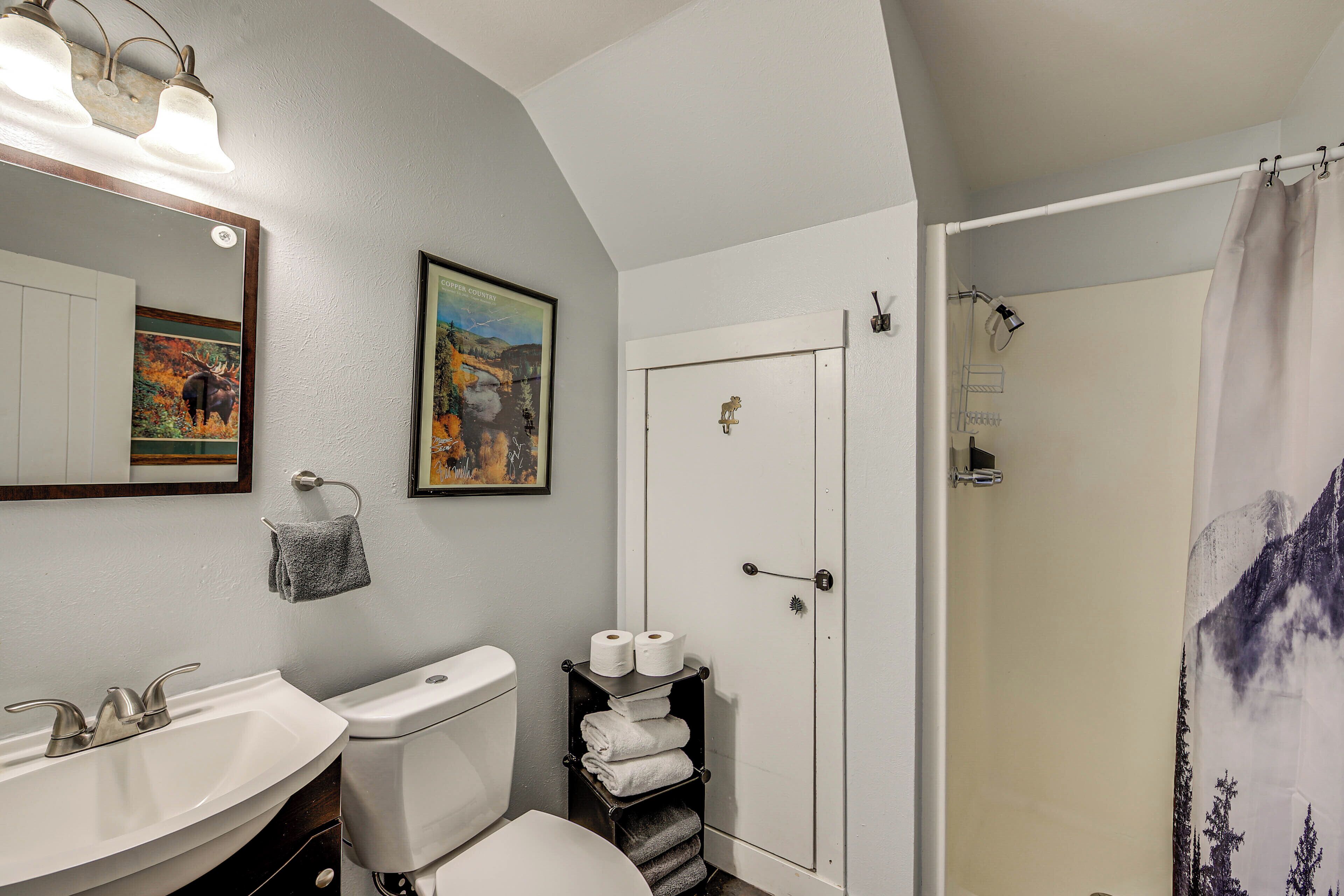 A bathroom with a sink, toilet, and shower. There are towels and toilet paper on a shelf.