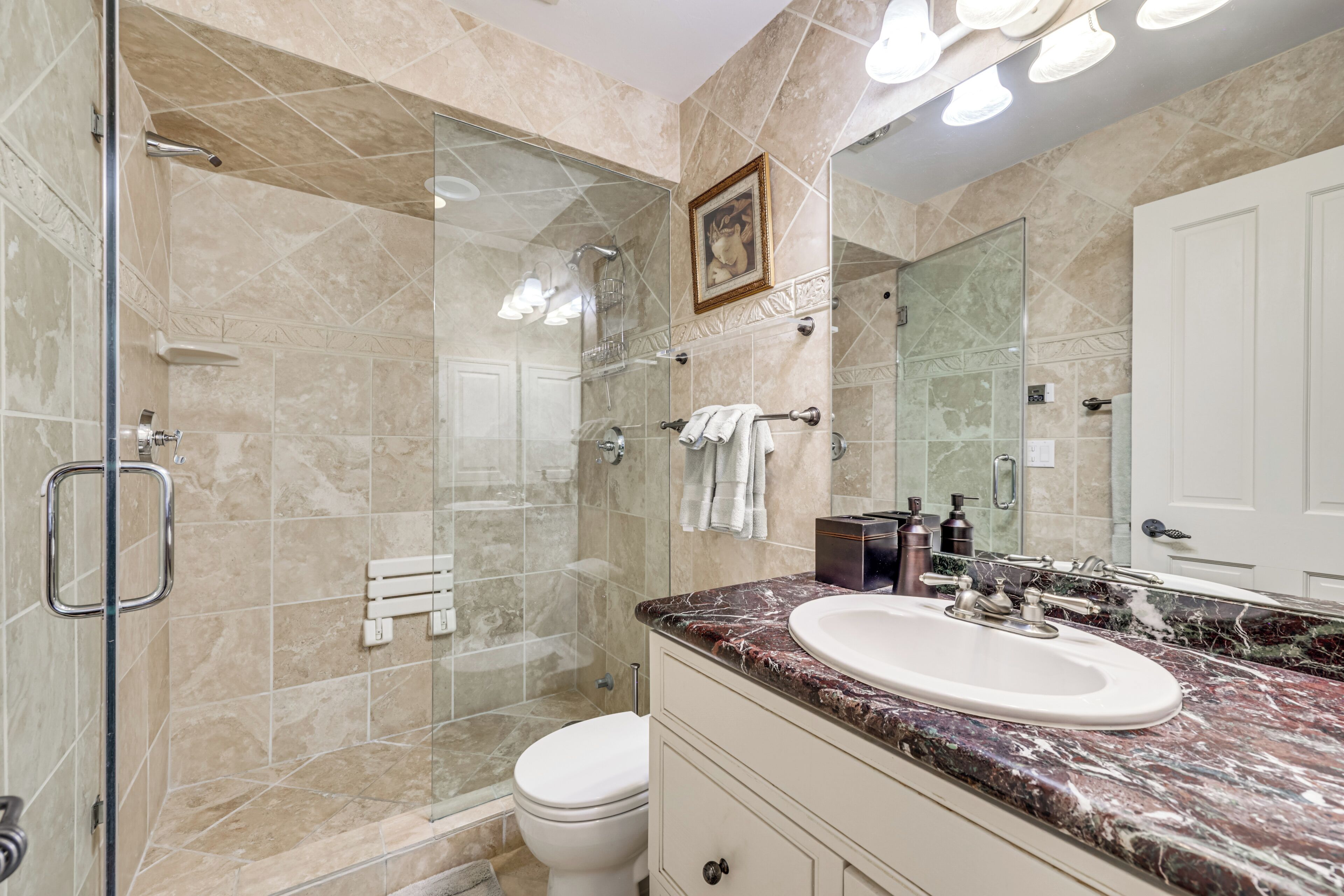 A bathroom with a glass shower enclosure, a white toilet, a sink with a marble countertop, and a large mirror.