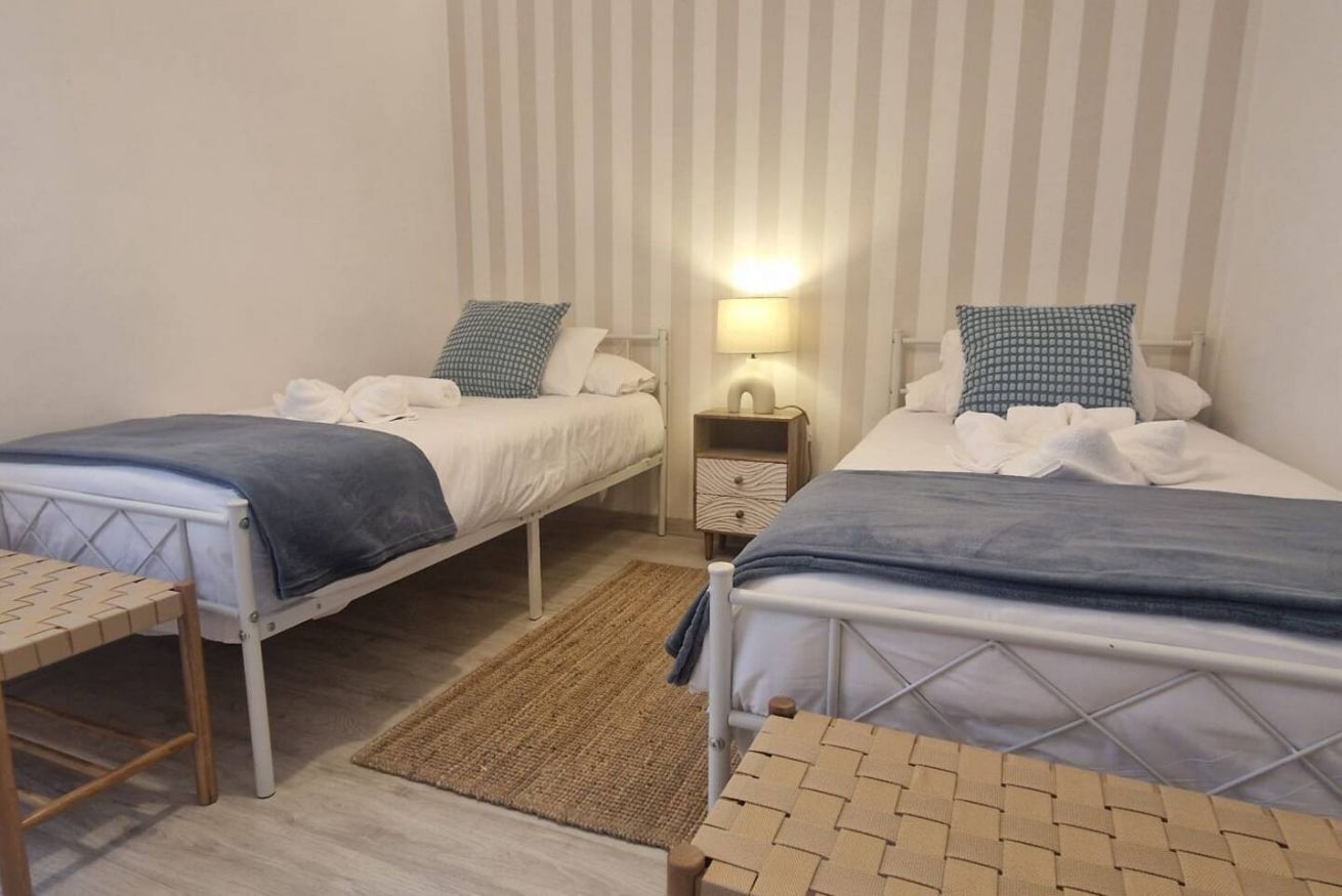 A room with two single beds, a small table, a nightstand with a lamp, and a woven rug.