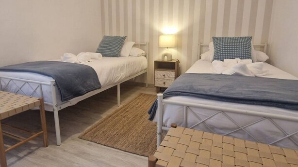 A room with two single beds, a small table, a nightstand with a lamp, and a woven rug. - 102B Poniente Mágico by R2R Consulting (Gijón)