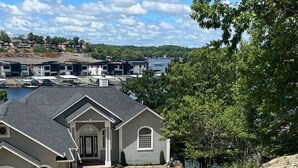 A residential area with a driveway leading to a house with a grey exterior and a black roof. There are multiple houses in the background, and a body of water is visible. - Lakefront property w dock & amazing sunsets  (Sunrise Beach)