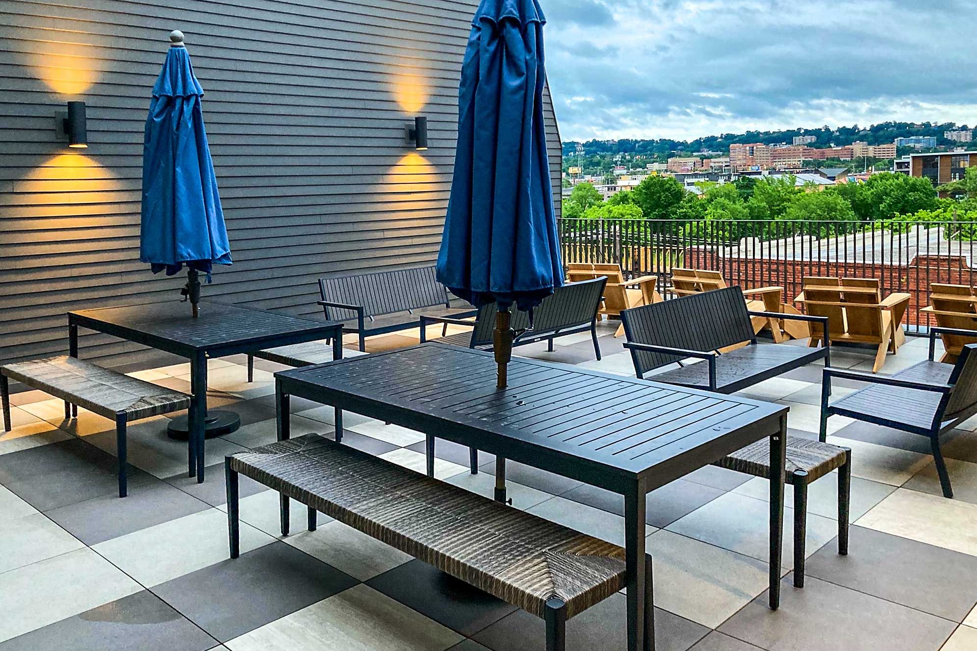 En udendørs terrasse med borde og stole, to blå parasoller og udsigt til bybilledet i baggrunden.