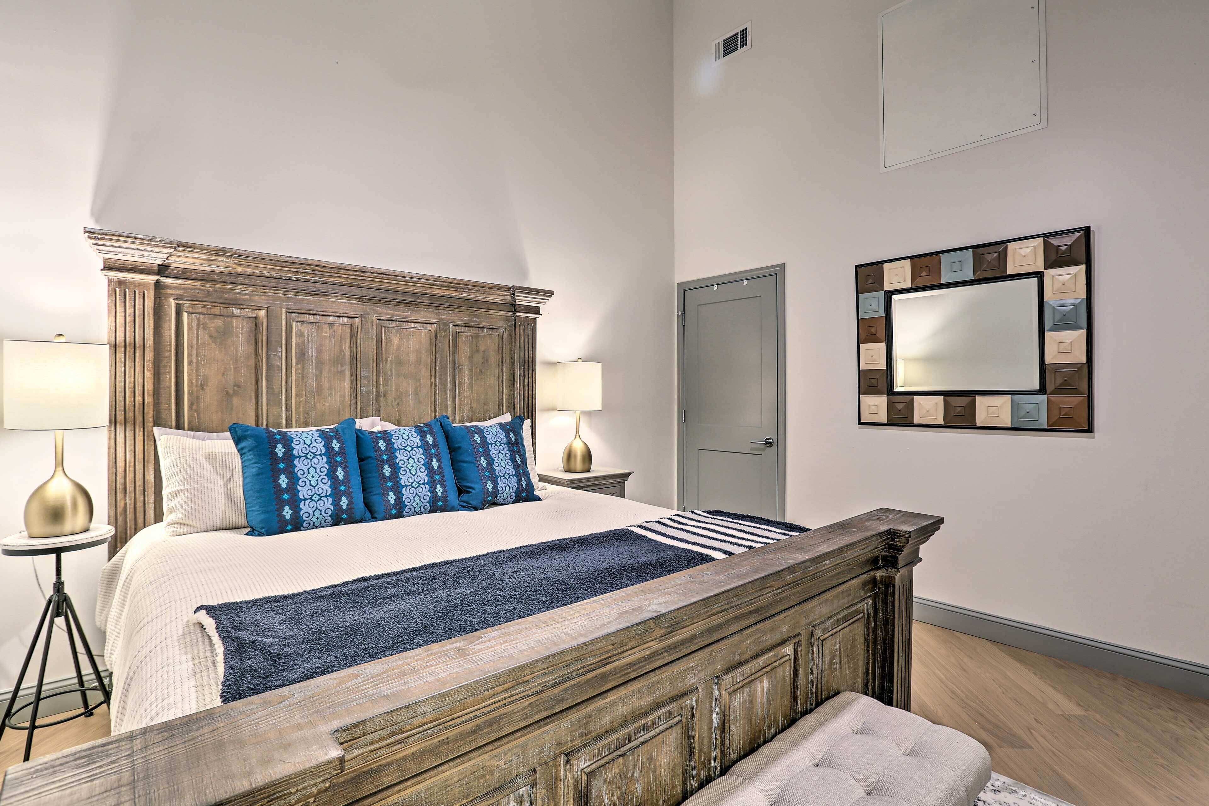 A bedroom with a wooden headboard, a large bed with patterned pillows, two bedside lamps, a bedside table, a door, and a wall-mounted mirror.