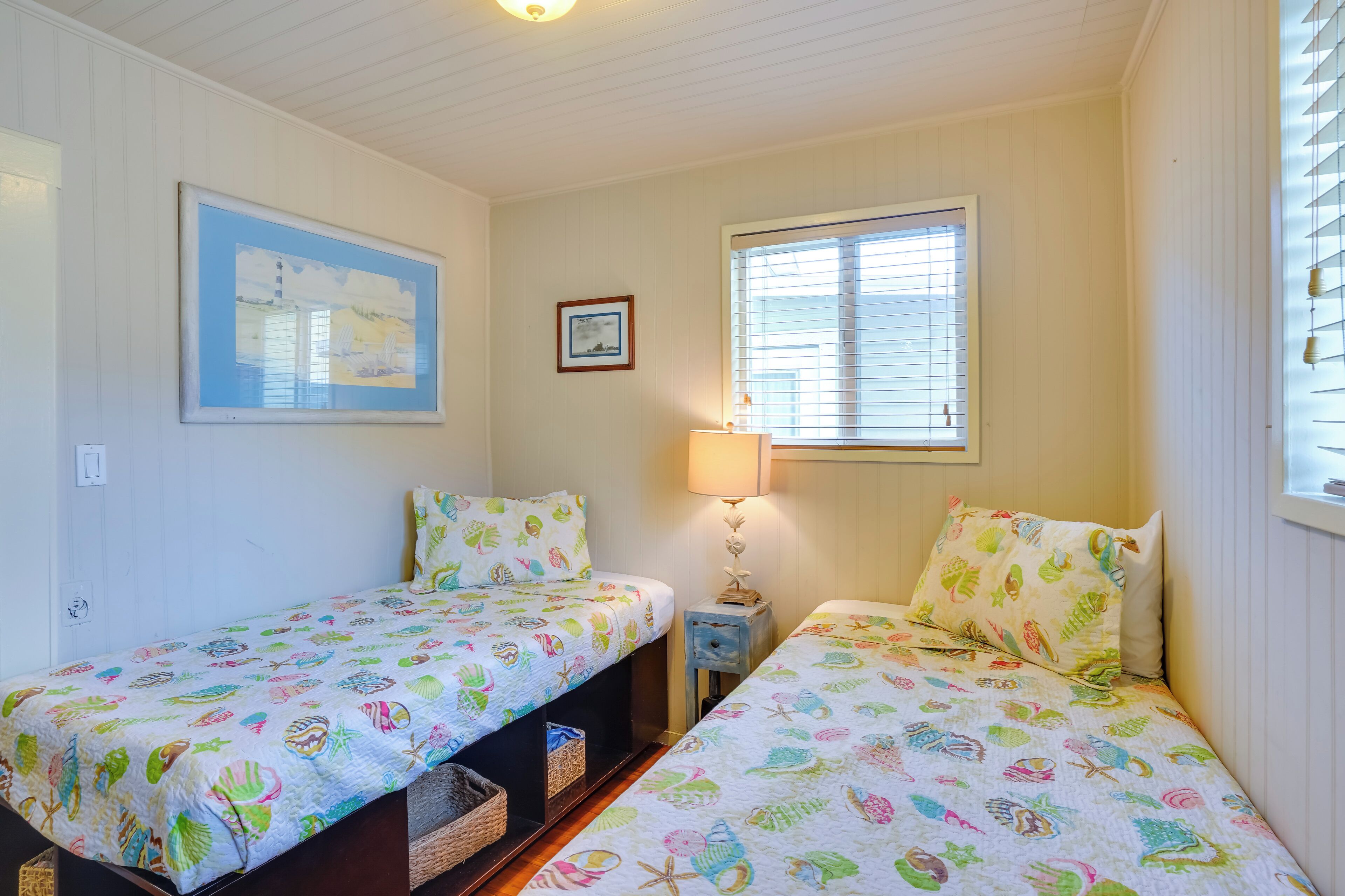A room with two single beds, each with a colorful bedspread featuring sea creatures. There is a small table with a lamp between the beds, and a window with blinds on the right wall.