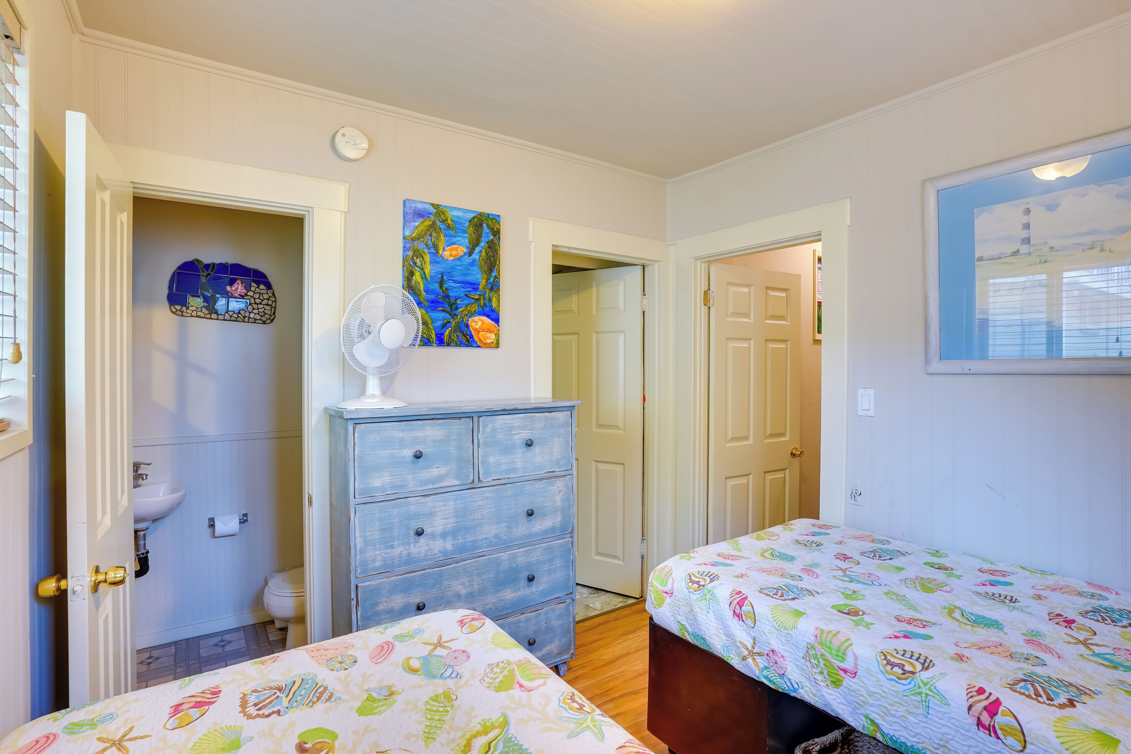 Une chambre avec deux lits, une commode, un ventilateur et une salle de bain visible à travers une porte ouverte.