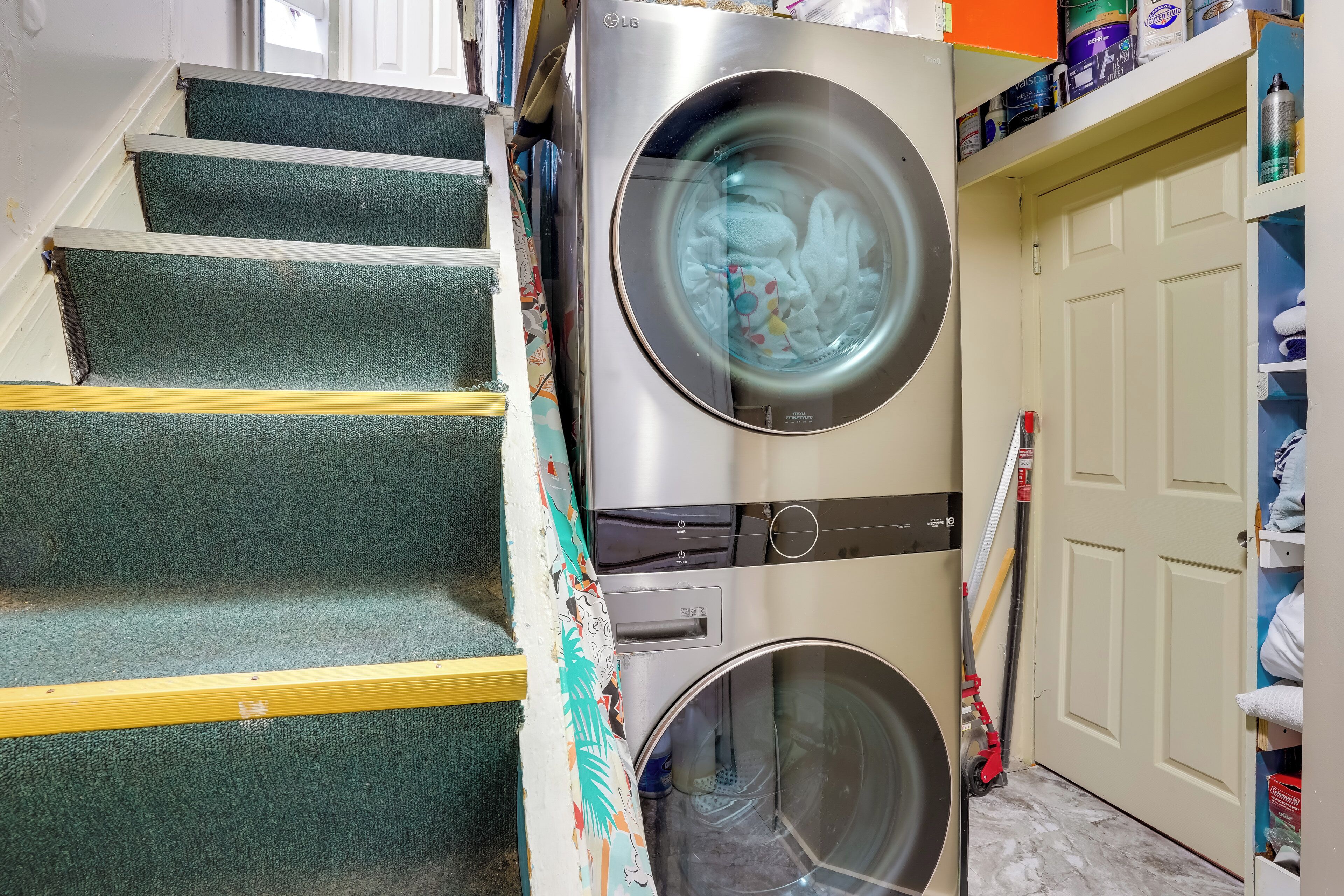 A laundry room with a front-loading washer and dryer, a broom, and a door leading to another room.