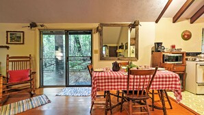 Un comedor con mantel a cuadros rojo y blanco, sillas de madera, una mesa de comedor de madera, una mecedora de madera, un cojín rojo, piso de madera, un gran ventanal corredizo que da a un balcón, una cocina con microondas, estufa y armarios, y un espejo en la pared.
