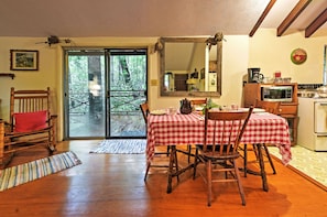 Un área de comedor con mantel a cuadros rojo y blanco, sillas de madera, una mesa de comedor de madera, una mecedora de madera, una zona de cocina con microondas y estufa, y un espejo grande en la pared.
