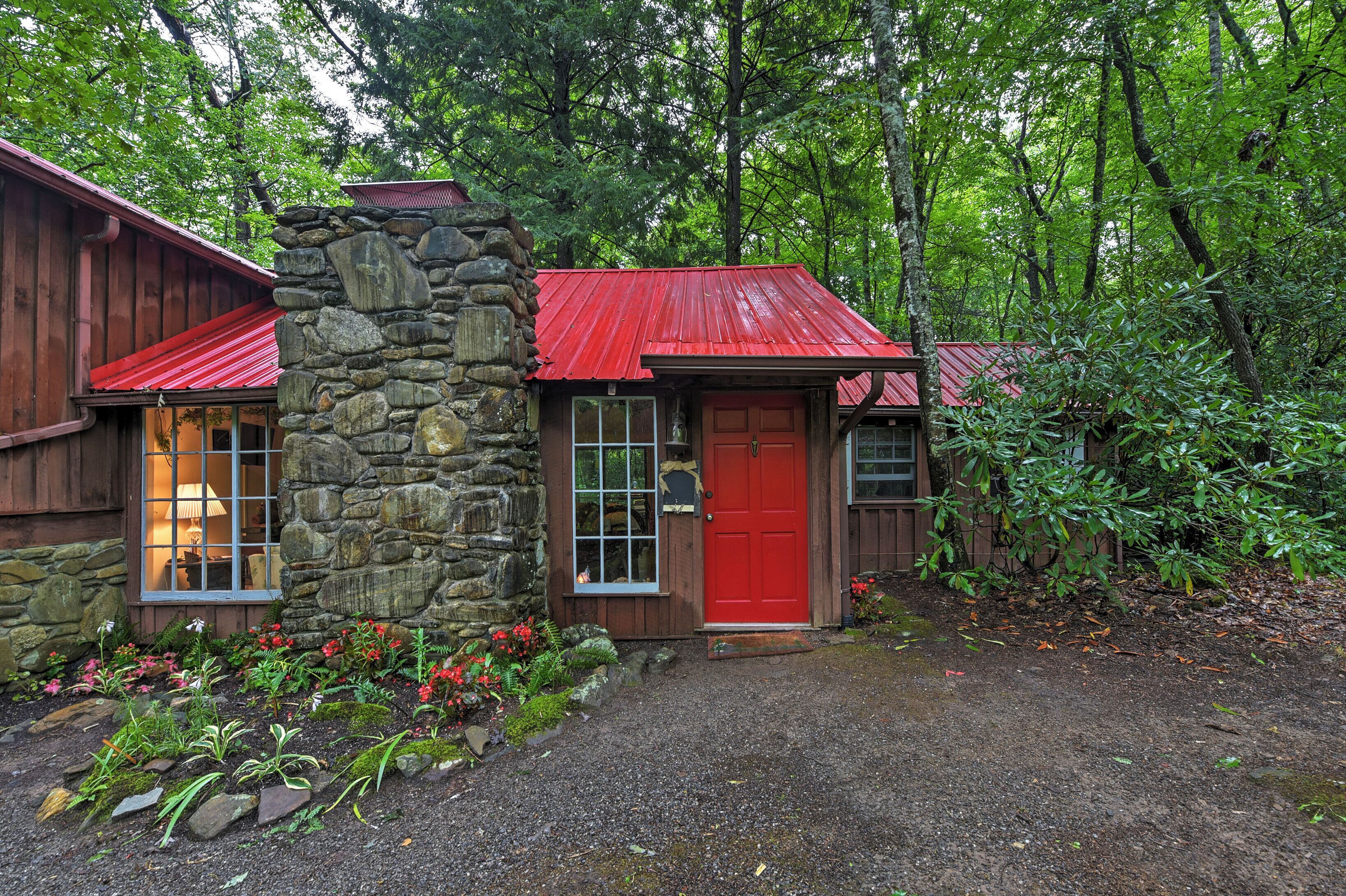 A rustic cabin with a stone chimney, a red door, and a gravel driveway surrounded by greenery.
