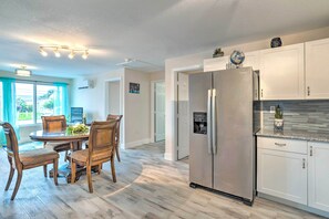 A modern kitchen with a stainless steel refrigerator, a dining area with wooden chairs, and a view of the outdoors through large windows.