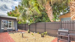 Un jardin avec un barbecue, une terrasse avec des chaises et un petit espace aménagé avec des plantes et un abreuvoir pour les oiseaux.