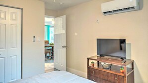 Une chambre à coucher avec un lit, un téléviseur posé sur un meuble en bois, un climatiseur et une porte donnant sur une autre pièce.