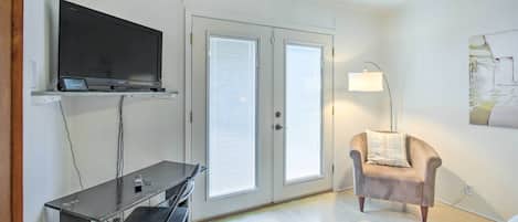 A room with a flat-screen TV on a stand, a desk with a chair, a lamp, and a chair with a pillow.