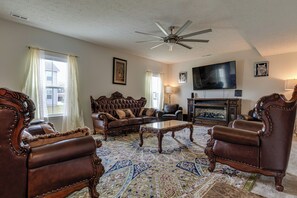 A spacious living room with a large ceiling fan, a fireplace, and a television mounted on the wall. - Spacious Home w/ Fireplace - Near Golf! (Brownsburg)