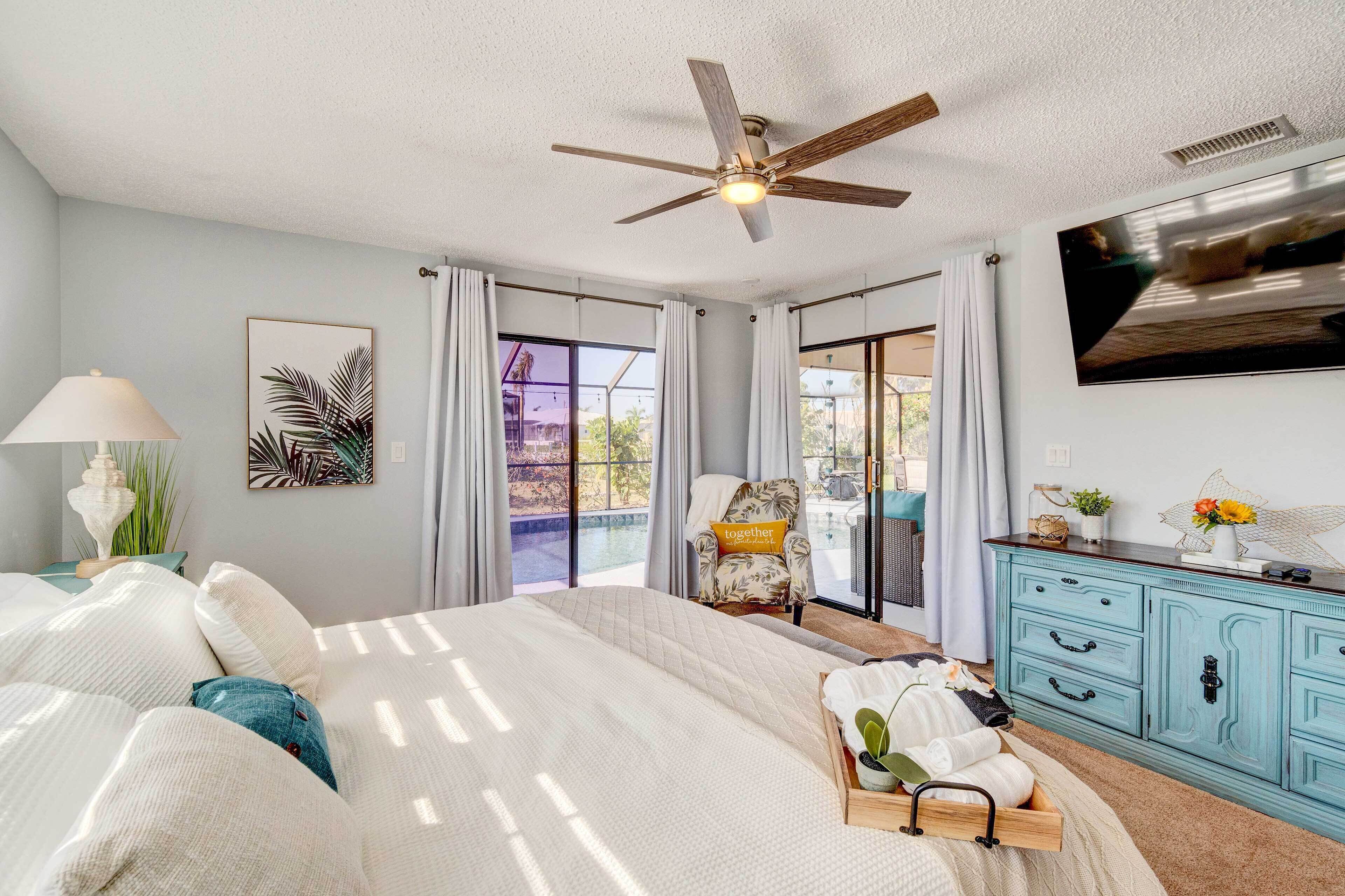 Une chambre à coucher comprenant un lit, un ventilateur de plafond, une télévision, une fenêtre avec des rideaux, une chaise et une commode.