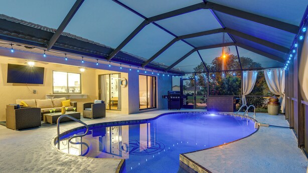 A covered outdoor swimming pool area with a curved design, a lounge with sofas and a coffee table, and a pergola with string lights. - Punta Gorda Canal Home w/ Private Pool! (Punta Gorda)