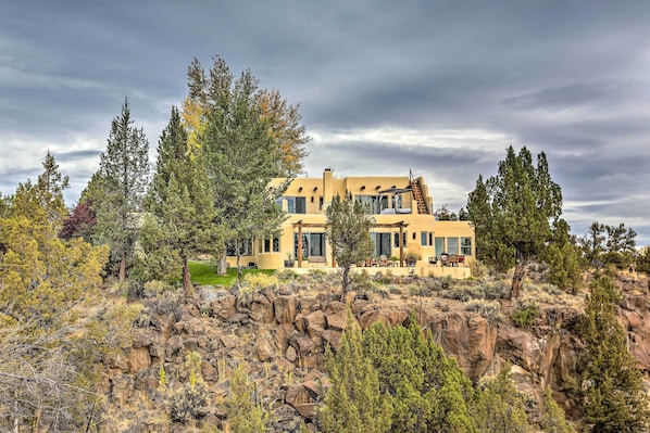 A modern two-story house with a flat roof, surrounded by a rocky landscape and sparse vegetation.