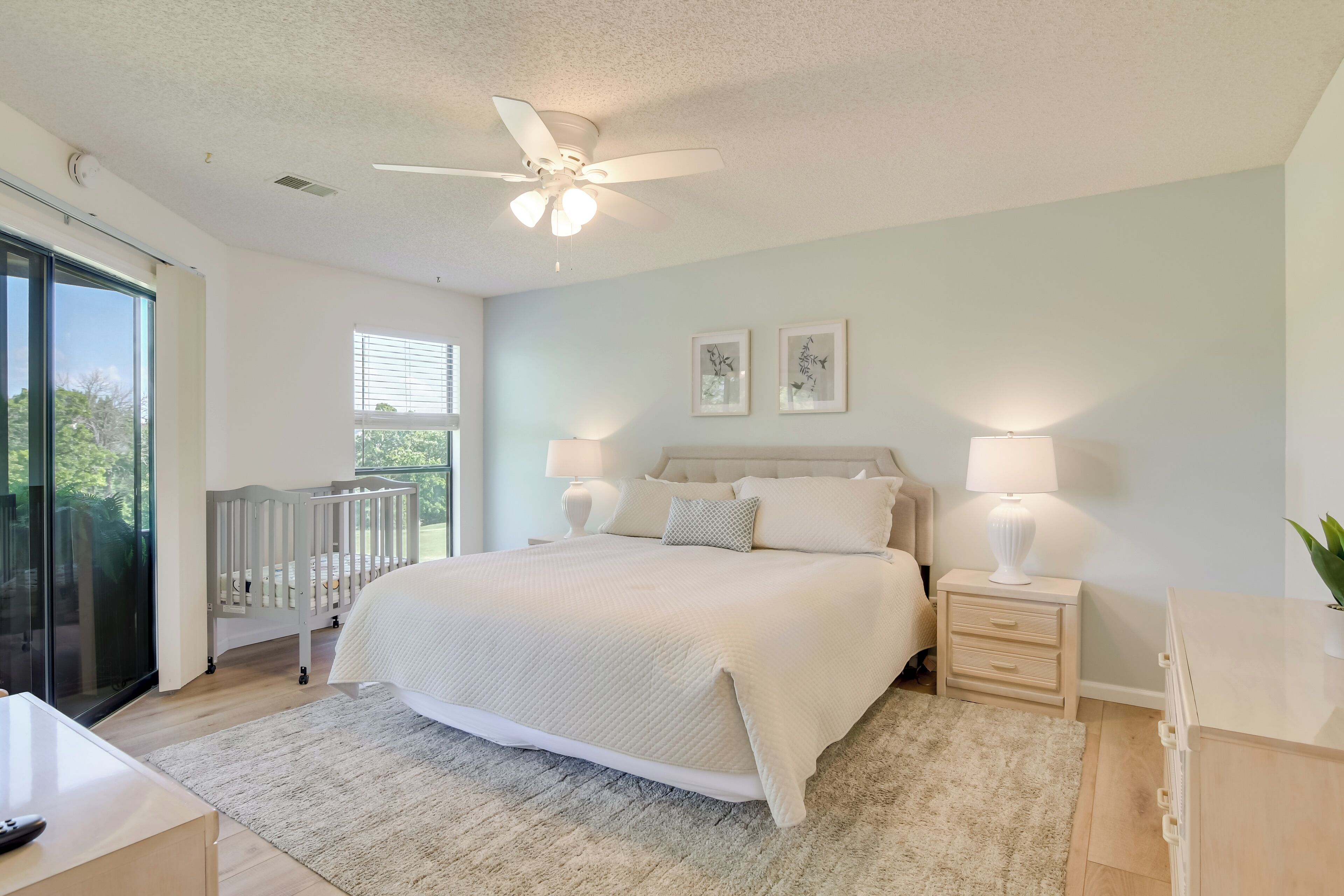 A bedroom with a large bed, two bedside tables with lamps, a ceiling fan, and a view through a sliding glass door.