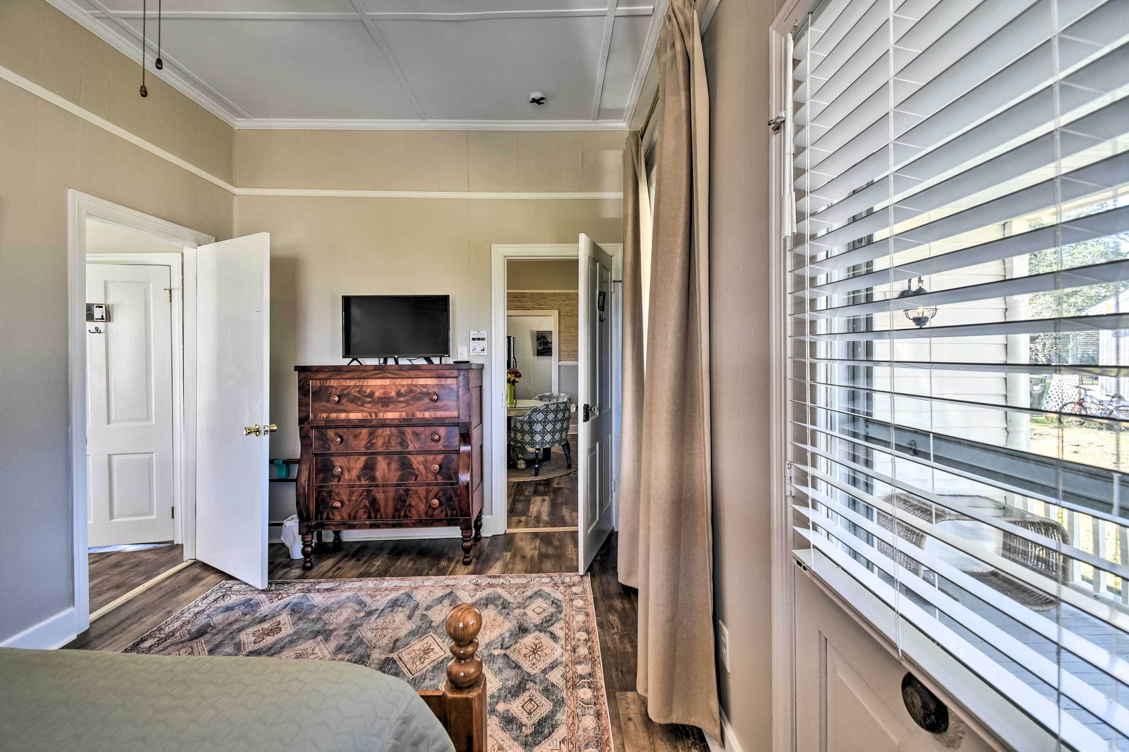 A hotel room with a wooden dresser, a television, a patterned rug, and a window with blinds.
