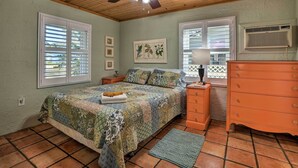 Ein Schlafzimmer mit Deckenventilator aus Holz, einem Bett mit farbenfroher Steppdecke, einem Schminktisch und einem Fenster mit Jalousien.