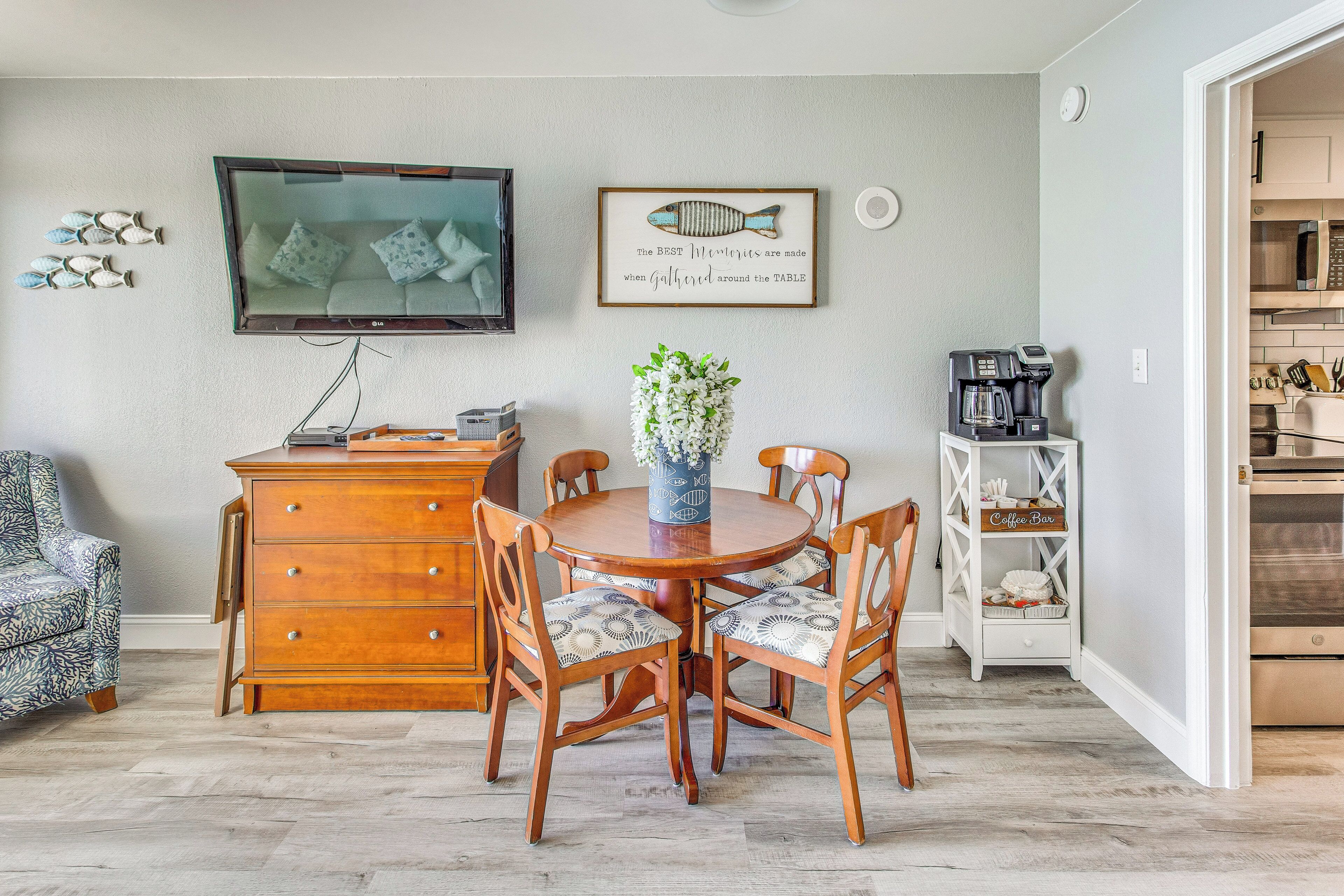 A dining area with a wooden table and chairs, a television mounted on the wall, and a coffee machine on a shelf.
