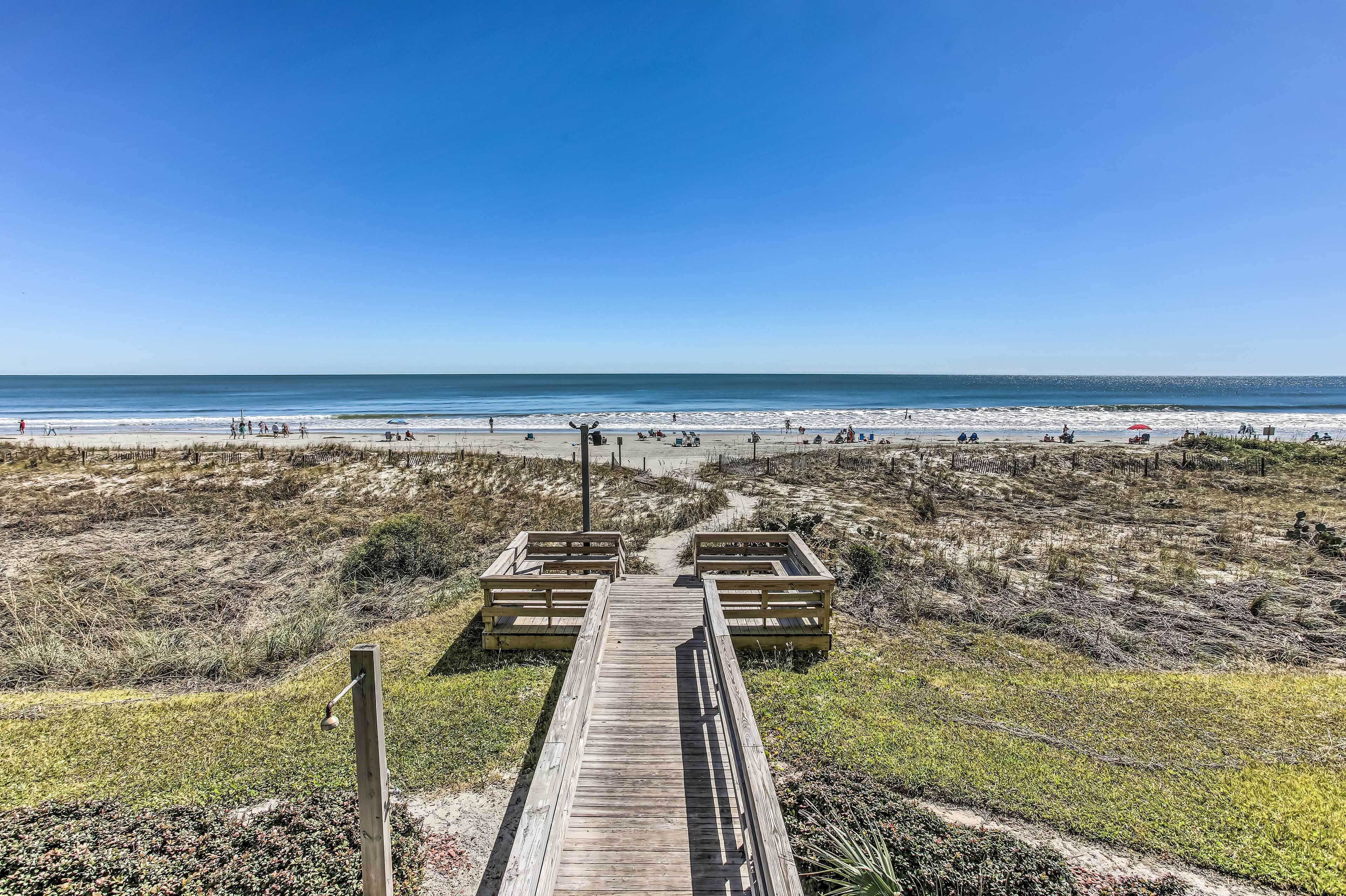 A wooden boardwalk leading to beach access points with a clear view of the ocean and beachgoers.