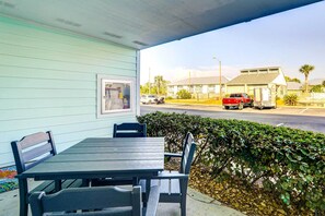 A covered outdoor seating area with a table and chairs, a framed picture on the wall, and a view of a street with a red car and a building in the background.
