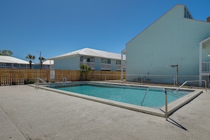 Una piscina con cerramiento de madera, un edificio con techo blanco y un cielo azul despejado.