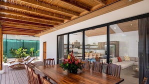 A spacious wooden deck with a dining table and chairs, surrounded by potted plants and a view of a swimming pool and a modern kitchen. - Casa Prema | Modern 3BR Home in Playa Grande (Cabo Velas)