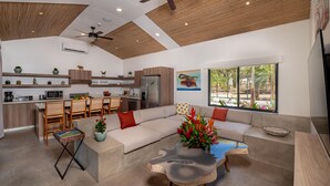 A modern kitchen with wooden cabinets, a built-in microwave, and a refrigerator. There is a dining area with a wooden table and chairs, and a living room with a sofa and a coffee table. - Casa Prema | Modern 3BR Home in Playa Grande (Cabo Velas)
